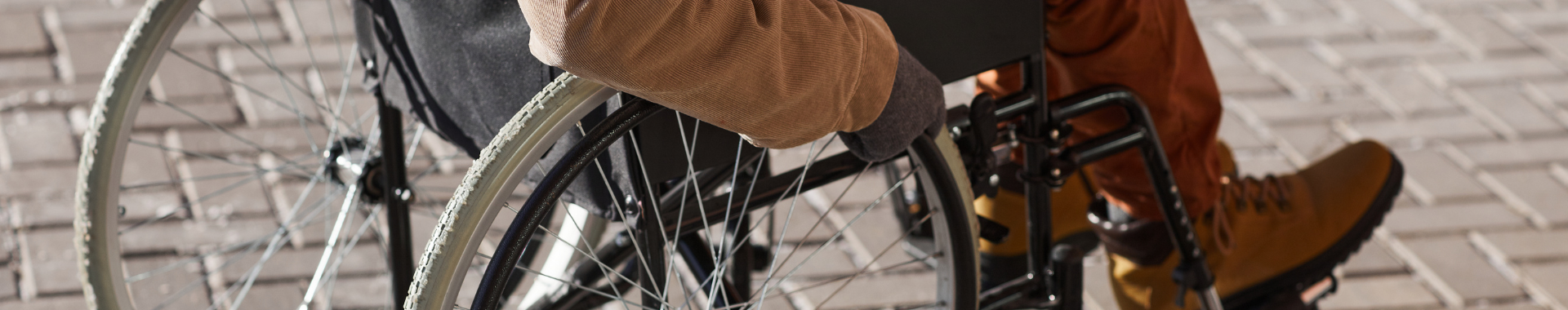 Das Bild zeigt eine Person in einem Rollstuhl. Der Fokus liegt auf dem rechten Arm und dem Rad des Rollstuhls. Die Person trägt braune Hosen und braune Schuhe. Der Boden besteht aus Pflastersteinen.