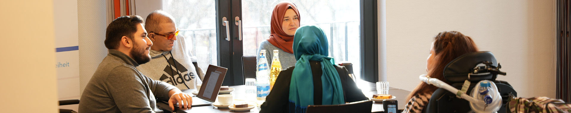 Das Bild zeigt fünf Personen an einem Tisch, die in einem Gespräch vertieft sind. Zwei der Frauen tragen ein Kopftuch, zwei Person sitzen in einem Rollstuhl. Auf dem Tisch stehen Getränke und Laptops.