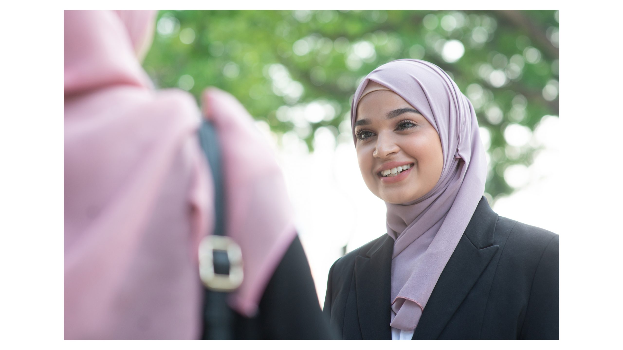 Zwei Muslimas schauen sich in unter dem Baum an. Die eine hat eine Business Blazer an mit einem helllila Kopftuch und die andere ein rosa Kopftuch.